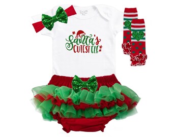Puede incluir: Un conjunto festivo para bebé con un body blanco con el texto "Santa's Cutest Elf", una diadema roja con un lazo de lentejuelas verdes, un tutú rojo y verde, un lazo de lentejuelas verdes y calcetines rojos y verdes.