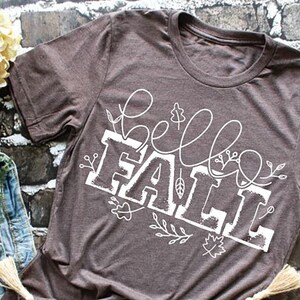 May include: A brown t-shirt with a white graphic that says "hello fall" with fall in a large font and hello in a smaller font. The graphic is surrounded by white floral designs.