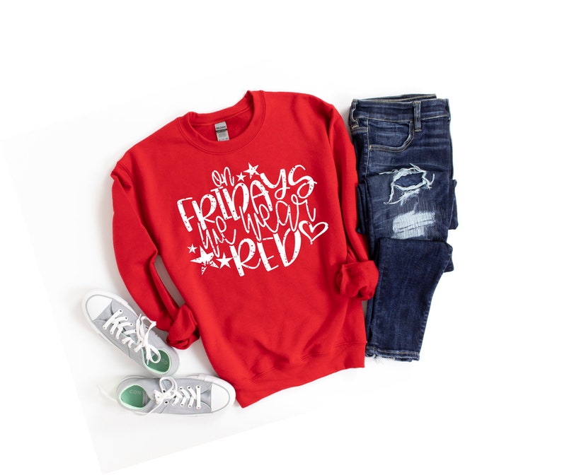K&ouml;nnte beinhalten: Roter Pullover mit dem Text "On Fridays we wear red" in Wei&szlig; mit Sternen und einem Herz. Der Pullover liegt flach auf einem wei&szlig;en Hintergrund mit einem Paar grauer Turnschuhe und einer blauen Jeans.