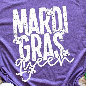 May include: Purple t-shirt with white text that reads "Mardi Gras queen" with fleur-de-lis designs.