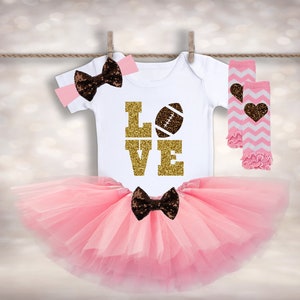 Puede incluir: Un tut&uacute; rosa, un body blanco con "LOVE" en purpurina dorada y una pelota de f&uacute;tbol marr&oacute;n, una diadema con lazo marr&oacute;n y polainas a rayas rosa y blancas con un coraz&oacute;n marr&oacute;n.