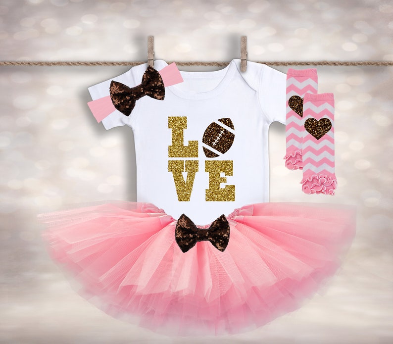 Puede incluir: Un tut&uacute; rosa, un body blanco con "LOVE" en purpurina dorada y una pelota de f&uacute;tbol marr&oacute;n, una diadema con lazo marr&oacute;n y polainas a rayas rosa y blancas con un coraz&oacute;n marr&oacute;n.