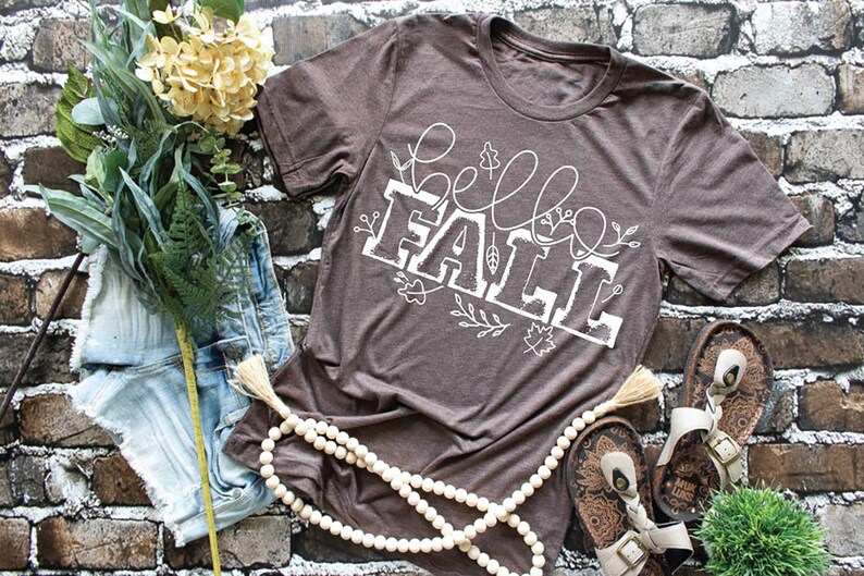 May include: A brown t-shirt with a white graphic that says "hello fall" with fall in a larger font and surrounded by small white floral designs.