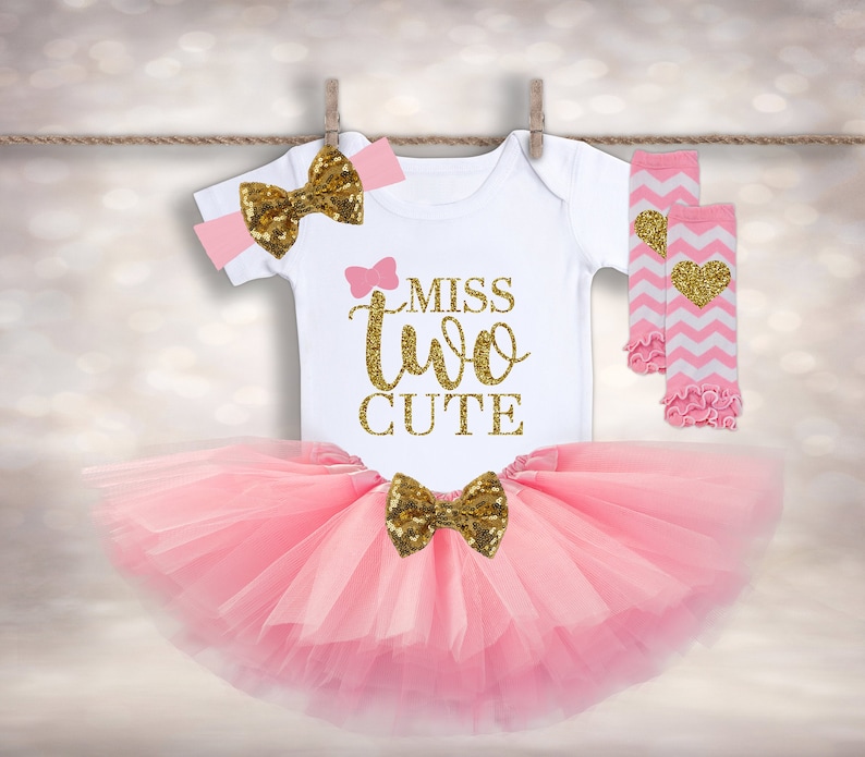 May include: A pink tutu with a gold glitter bow, a white onesie with the text "Miss Two Cute" in gold glitter, a pink headband with a gold glitter bow, and pink and white leg warmers with a gold glitter heart.