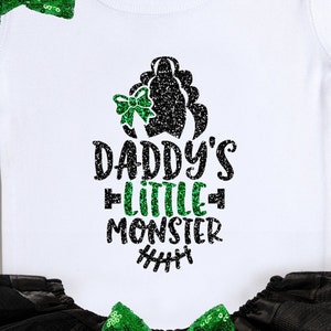 Op de afbeelding: Witte romper met een zwart en groen glitterontwerp met de tekst "Daddy's Little Monster" met een groene strik en een zwart en groen glittermonsterhoofd.