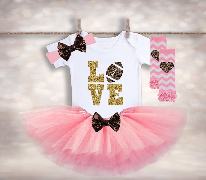 Puede incluir: Un tut&uacute; rosa, un body blanco con "LOVE" en purpurina dorada y un gr&aacute;fico de f&uacute;tbol americano marr&oacute;n, una diadema con lazo marr&oacute;n y polainas a rayas rosa y blancas con detalles de coraz&oacute;n marr&oacute;n.