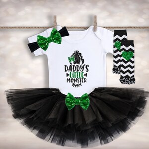 Op de afbeelding: Een zwarte tule tutu rok met een groen en zwart strikje, een wit shirt met de tekst "Daddy's Little Monster" in groene glitter en zwart-wit gestreepte beenwarmers met een groen glitter hart.