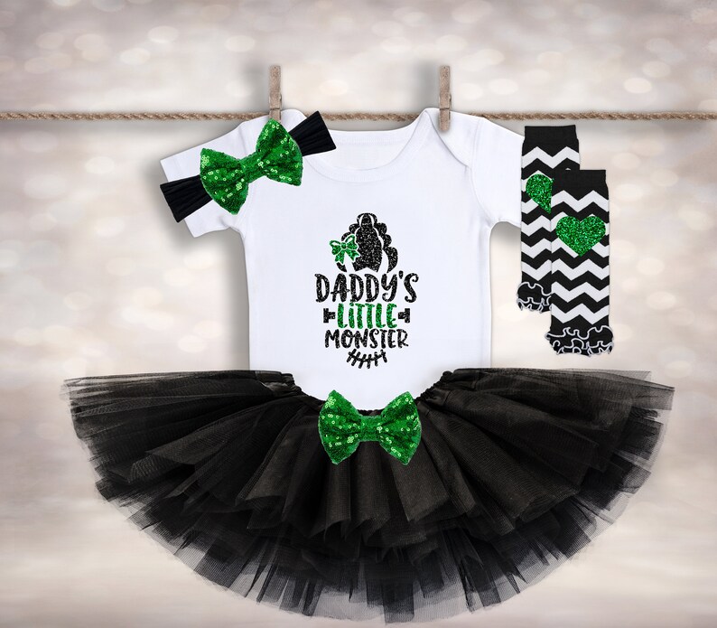Op de afbeelding: Een zwarte tule tutu rok met een groen en zwart strikje, een wit shirt met de tekst "Daddy's Little Monster" in groene glitter en zwart-wit gestreepte beenwarmers met een groen glitter hart.