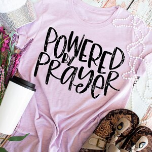 May include: A light pink t-shirt with the text "Powered by Prayer" in black distressed lettering. The shirt is laid flat on a wooden surface with a white mug, a string of pearls, and a pair of sandals.