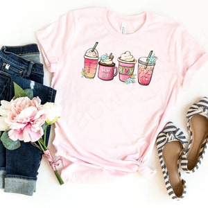 Op de afbeelding: Roze T-shirt met een afbeelding van vier verschillende koffiedrankjes. De drankjes zijn versierd met bloemen en de tekst "Mom Fuel". Het shirt wordt gecombineerd met een blauwe jeans, een boeket roze bloemen en gestreepte ballerina's.