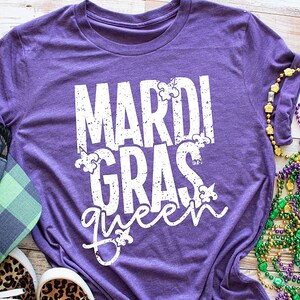 May include: Purple t-shirt with the text "Mardi Gras queen" in white with a distressed look. The shirt is surrounded by Mardi Gras beads and a green and black plaid hat.