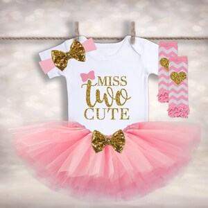 May include: Pink tutu with a gold bow, a white onesie with the text "Miss Two Cute" in gold glitter, a pink headband with a gold bow, and pink and white leg warmers with a gold heart.