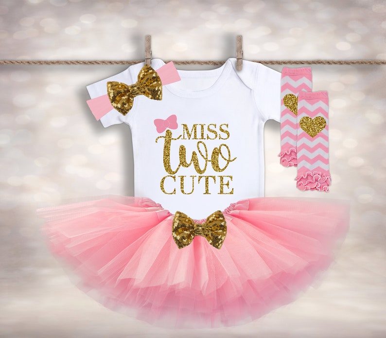 May include: Pink tutu with a gold bow, a white onesie with the text "Miss Two Cute" in gold glitter, a pink headband with a gold bow, and pink and white leg warmers with a gold heart.