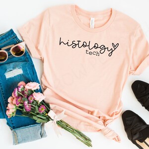 May include: A peach-colored t-shirt with the text "histology tech" and a small heart graphic printed on the front.