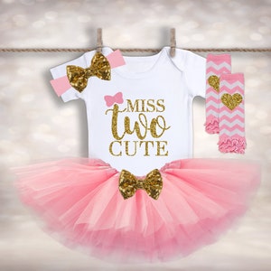 May include: A pink tutu with a gold bow, a white onesie with the text "Miss Two Cute" in gold glitter, a pink headband with a gold bow, and pink and white leg warmers with a gold heart.