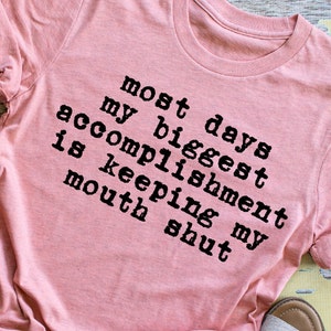 May include: Pink t-shirt with black text that reads "Most days my biggest accomplishment is keeping my mouth shut."