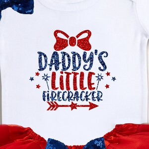May include: White baby onesie with red and blue glitter text that says "Daddy's Little Firecracker" with a red glitter bow and a red and blue glitter arrow. The onesie is paired with a red tulle skirt and a blue sequin bow.