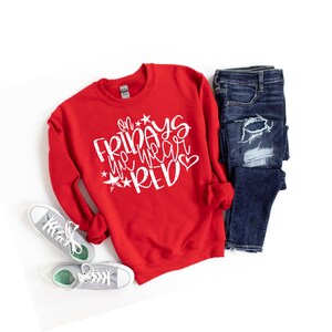 K&ouml;nnte beinhalten: Roter Pullover mit dem Text "On Fridays we wear red" in Wei&szlig; mit Sternen und einem Herz. Der Pullover liegt flach auf einem wei&szlig;en Hintergrund mit einem Paar grauer Turnschuhe und einer blauen Jeans.