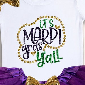 May include: White bodysuit with a gold and green glitter design that says "It's Mardi Gras y'all". The bodysuit is paired with a purple tulle skirt and a gold bow.