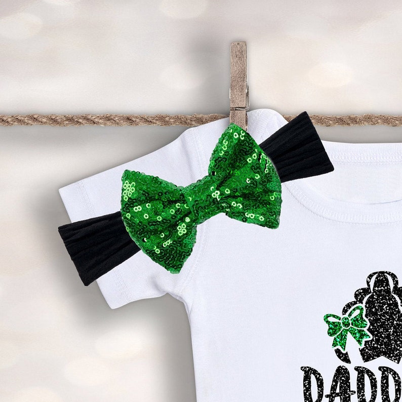 Op de afbeelding: Een wit shirt met een zwart en groen pailletten haarband. Het shirt heeft een zwart silhouet van een meisje met een strik in haar haar en de tekst "Daddy's Girl".