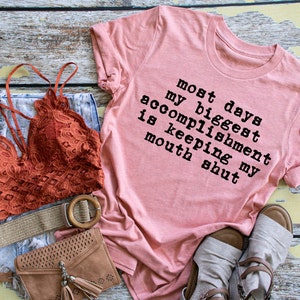May include: A pink t-shirt with the text "most days my biggest accomplishment is keeping my mouth shut" printed on it.