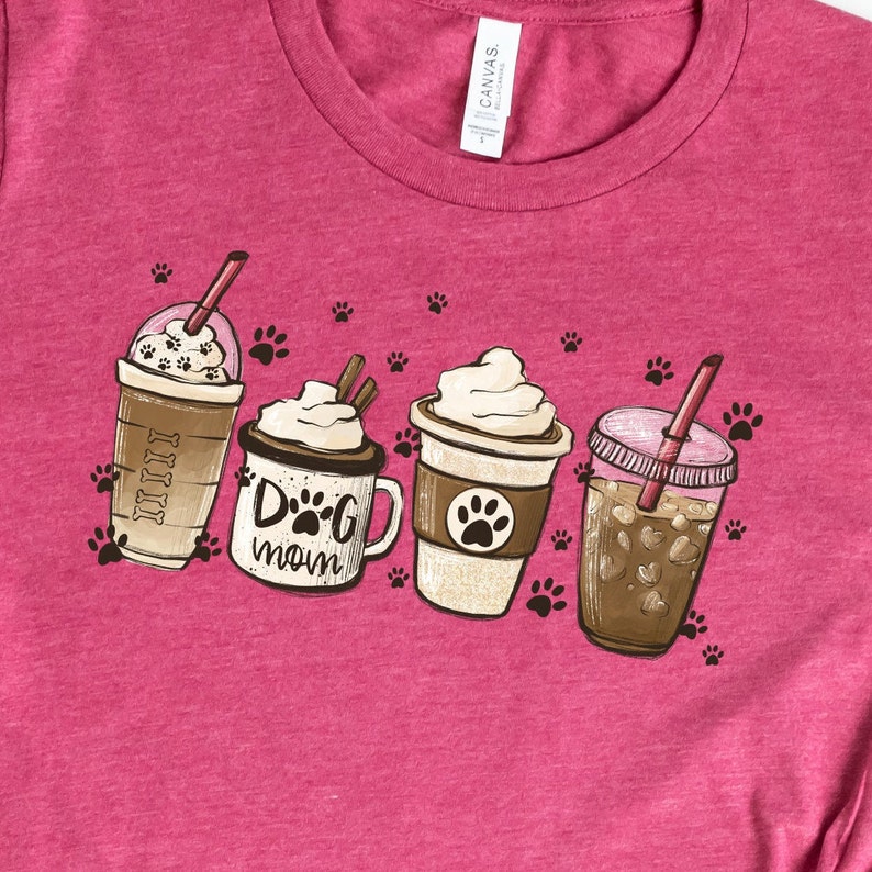 Pode incluir: Uma camiseta rosa com um gr&aacute;fico de quatro bebidas de caf&eacute;. As bebidas s&atilde;o um caf&eacute; gelado, um chocolate quente, um latte e um caf&eacute; gelado com chantilly. O gr&aacute;fico tamb&eacute;m inclui pegadas.