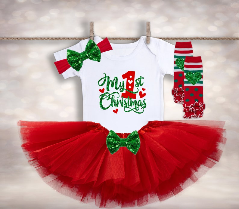 May include: A red tulle tutu skirt with a green and white bow, a white onesie with the text "My 1st Christmas" in red and green glitter, and a pair of red and green striped leg warmers with a green heart design.