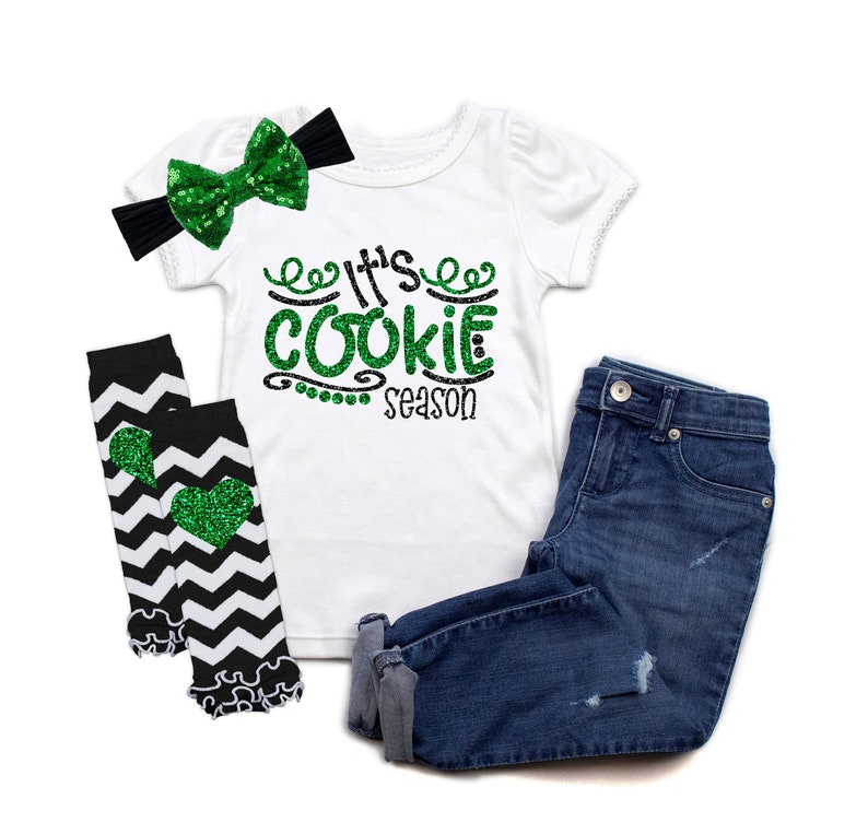 May include: A white t-shirt with green glitter text that says "It's Cookie Season". The shirt is paired with black and white chevron leg warmers with green glitter hearts and a black headband with a green glitter bow.