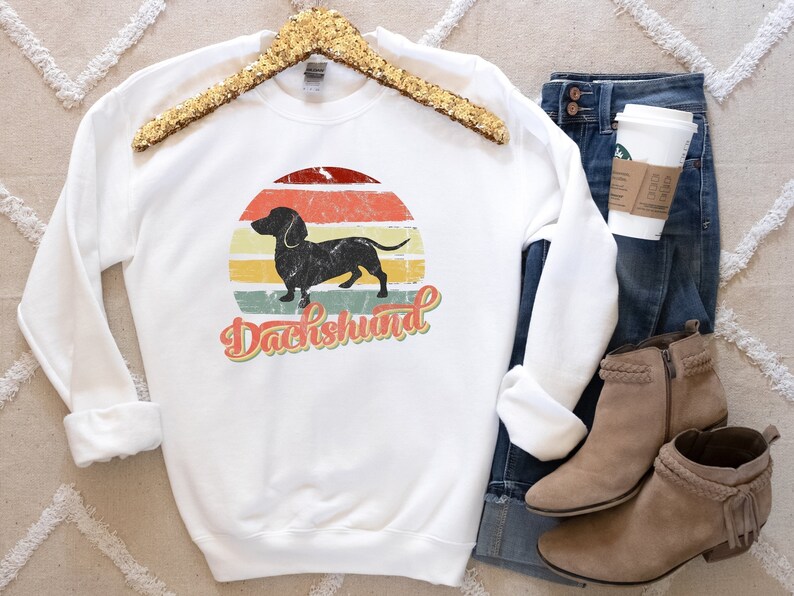May include: White sweatshirt with a vintage-style graphic of a dachshund silhouette in front of a sunset with the word "Dachshund" in a retro font.