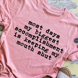 May include: A pink t-shirt with the text "Most days my biggest accomplishment is keeping my mouth shut" printed in black.