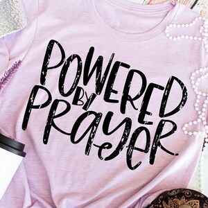 May include: A pink t-shirt with the text "Powered by Prayer" in black distressed lettering.