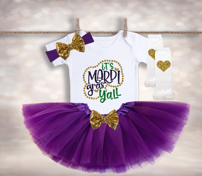 May include: Purple tulle tutu with a gold sequin bow, a white bodysuit with the text "It's Mardi Gras Y'all" in gold and green glitter, and white leg warmers with a gold glitter heart.