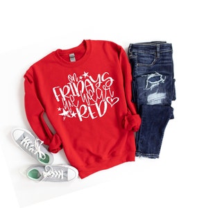 K&ouml;nnte beinhalten: Roter Pullover mit wei&szlig;em Text, der "On Fridays we wear red" mit Sternen und einem Herzen sagt. Der Pullover liegt auf einem wei&szlig;en Hintergrund mit einer Jeans und wei&szlig;en Turnschuhen.