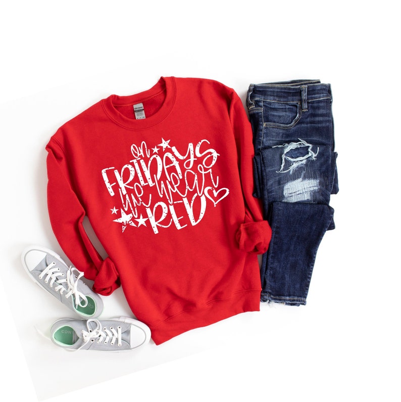 K&ouml;nnte beinhalten: Roter Pullover mit wei&szlig;em Text, der "On Fridays we wear red" mit Sternen und einem Herzen sagt. Der Pullover liegt auf einem wei&szlig;en Hintergrund mit einer Jeans und wei&szlig;en Turnschuhen.