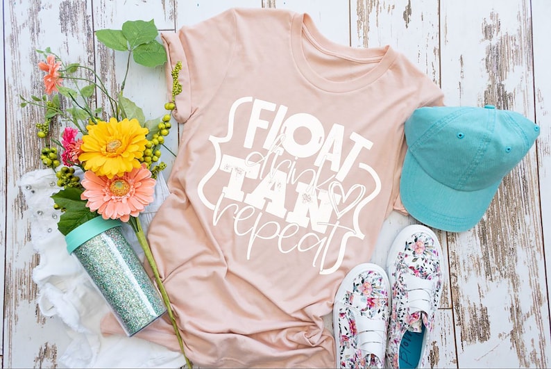 以下が含まれることがあります： 白い文字で「FLOAT drink TAN repeat」と書かれたピンクのTシャツ。白い木製の表面に、ターコイズブルーの野球帽、花柄の靴、キラキラ輝く透明なタンブラーと一緒に置かれています。