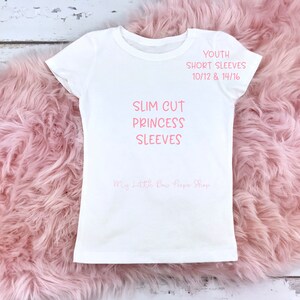 May include: A white youth t-shirt with short sleeves. The shirt features the text "SLIM CUT PRINCESS SLEEVES" and "YOUTH SHORT SLEEVES 10/12 & 14/16" in pink. The shop name "My Little Bow Peeps Shop" is also visible. The shirt is displayed on a pink, fluffy surface.