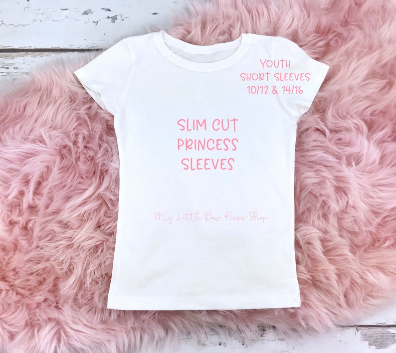 May include: A white youth t-shirt with short sleeves. The shirt features the text "SLIM CUT PRINCESS SLEEVES" and "YOUTH SHORT SLEEVES 10/12 & 14/16" in pink. The shop name "My Little Bow Peeps Shop" is also visible. The shirt is displayed on a pink, fluffy surface.
