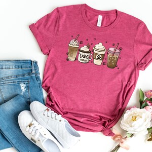 Pode incluir: Uma camiseta rosa com um gr&aacute;fico de quatro x&iacute;caras de caf&eacute; com chantilly e um canudo. O texto em uma das x&iacute;caras diz "Dog Mom". A camiseta est&aacute; cercada por jeans, t&ecirc;nis brancos e flores cor-de-rosa.