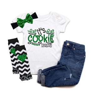 May include: A white t-shirt with green glitter text that says "It's Cookie Season". The shirt is paired with black and white chevron leg warmers with green glitter hearts, a black headband with a green glitter bow, and blue denim jeans.