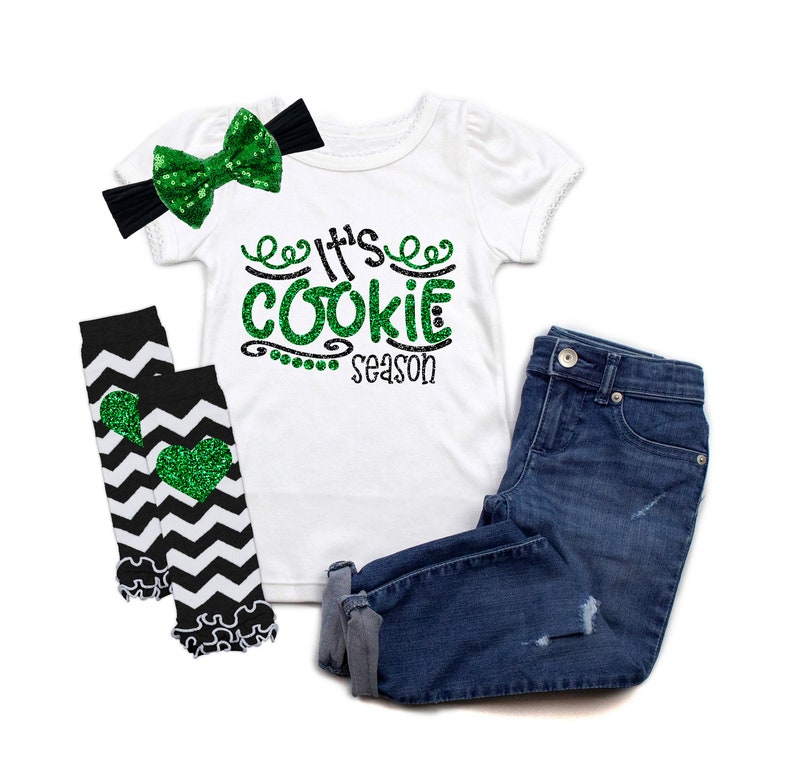 May include: A white t-shirt with green glitter text that says "It's Cookie Season". The shirt is paired with black and white chevron leg warmers with green glitter hearts, a black headband with a green glitter bow, and blue denim jeans.