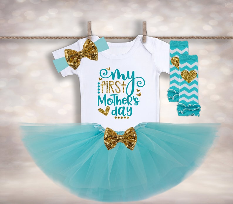 Puede incluir: Un conjunto para beb&eacute; ni&ntilde;a, que incluye un body blanco con el texto "My First Mother's Day", una falda tut&uacute; turquesa, una diadema y calentadores de piernas. El conjunto presenta lazos de lentejuelas doradas y detalles de coraz&oacute;n. Los calentadores tienen un patr&oacute;n de chevron.