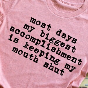 May include: Pink t-shirt with black text that reads "Most days my biggest accomplishment is keeping my mouth shut."