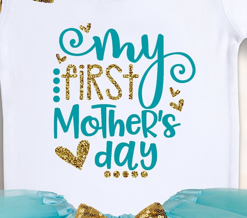 Puede incluir: Body de beb&eacute; blanco con el texto "My First Mother's Day" en verde azulado y purpurina dorada. El dise&ntilde;o incluye corazones y puntos. Un tut&uacute; azul claro con lentejuelas doradas es visible en la parte inferior.