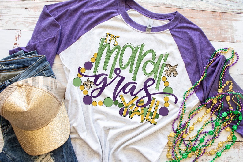 May include: A white and purple raglan t-shirt with the text "It's Mardi Gras Y'all" in green and gold lettering. The shirt is decorated with green, gold, and purple beads and a fleur-de-lis.