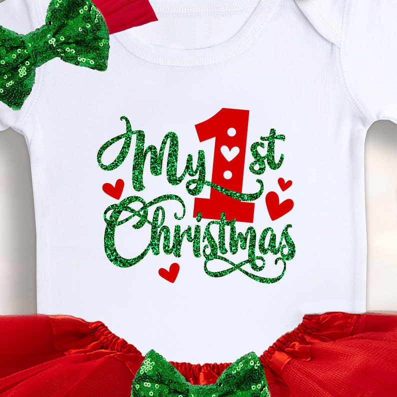 May include: A white onesie with a red number one and the text "My 1st Christmas" in green glitter. The onesie is paired with a red tulle skirt and a green sequin bow.