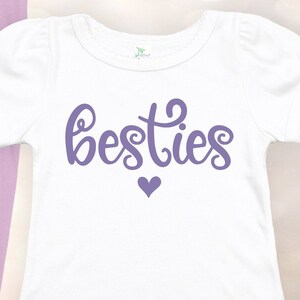 K&ouml;nnte beinhalten: Wei&szlig;es T-Shirt mit lilafarbenem Text "besties" und einem kleinen lilafarbenen Herz.
