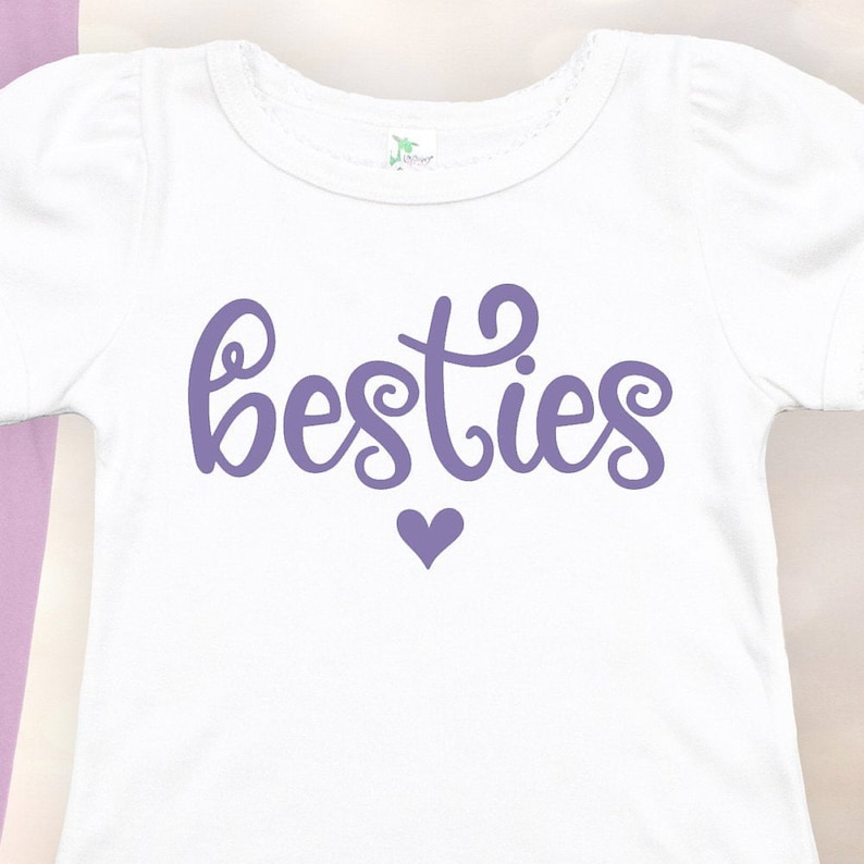 K&ouml;nnte beinhalten: Wei&szlig;es T-Shirt mit lilafarbenem Text "besties" und einem kleinen lilafarbenen Herz.