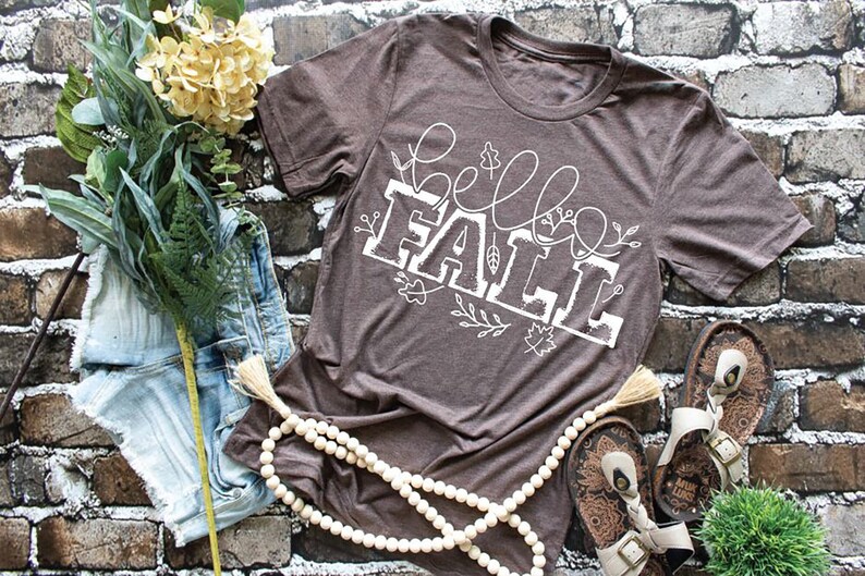 May include: A brown t-shirt with a white graphic that says "hello fall" with fall in a large font and hello in a smaller font. The graphic is surrounded by white floral designs.