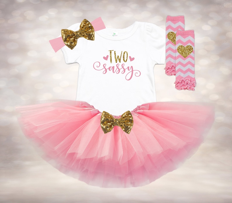 May include: Pink tulle tutu skirt with a gold glitter bow, a white t-shirt with the text "TWO sassy" in pink, a pink headband with a gold glitter bow, and white and pink leg warmers with a gold heart.