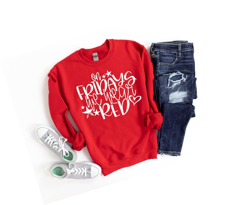 K&ouml;nnte beinhalten: Roter Pullover mit wei&szlig;em Text, der "On Fridays we wear red" mit Sternen und einem Herz sagt. Der Pullover liegt auf einer wei&szlig;en Oberfl&auml;che mit einem Paar grauer Turnschuhe und einer blauen Jeans.